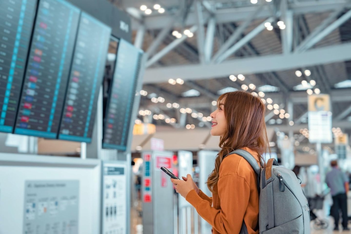 공항 출국장에서 배낭을 메고 스마트폰을 든 여성이 비행기 시간표가 적힌 대형 전광판을 확인하며 밝게 웃고 있는 모습입니다.