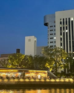 어스름한 저녁 하늘을 배경으로 강가에 위치한 화려한 조명의 테라스 카페와 현대적인 고층 빌딩들이 어우러진 평화로운 도시 야경입니다.