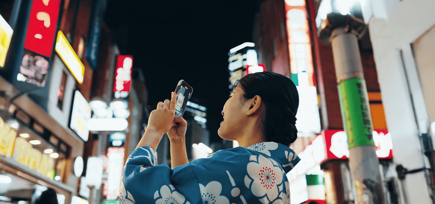 화려한 조명이 빛나는 일본 도심의 밤거리에서 푸른색 꽃무늬 유카타를 입은 여성이 스마트폰을 높이 들어 야경 사진을 찍고 있는 모습입니다.