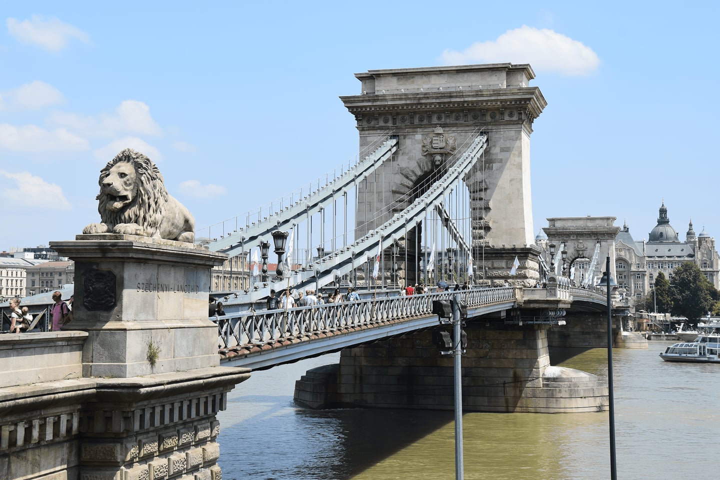 부다페스트 다뉴브 강의 상징적인 세체니 다리 입구에 세워진 위엄 있는 사자 석상과 웅장한 석조 주탑이 보이는 풍경입니다.