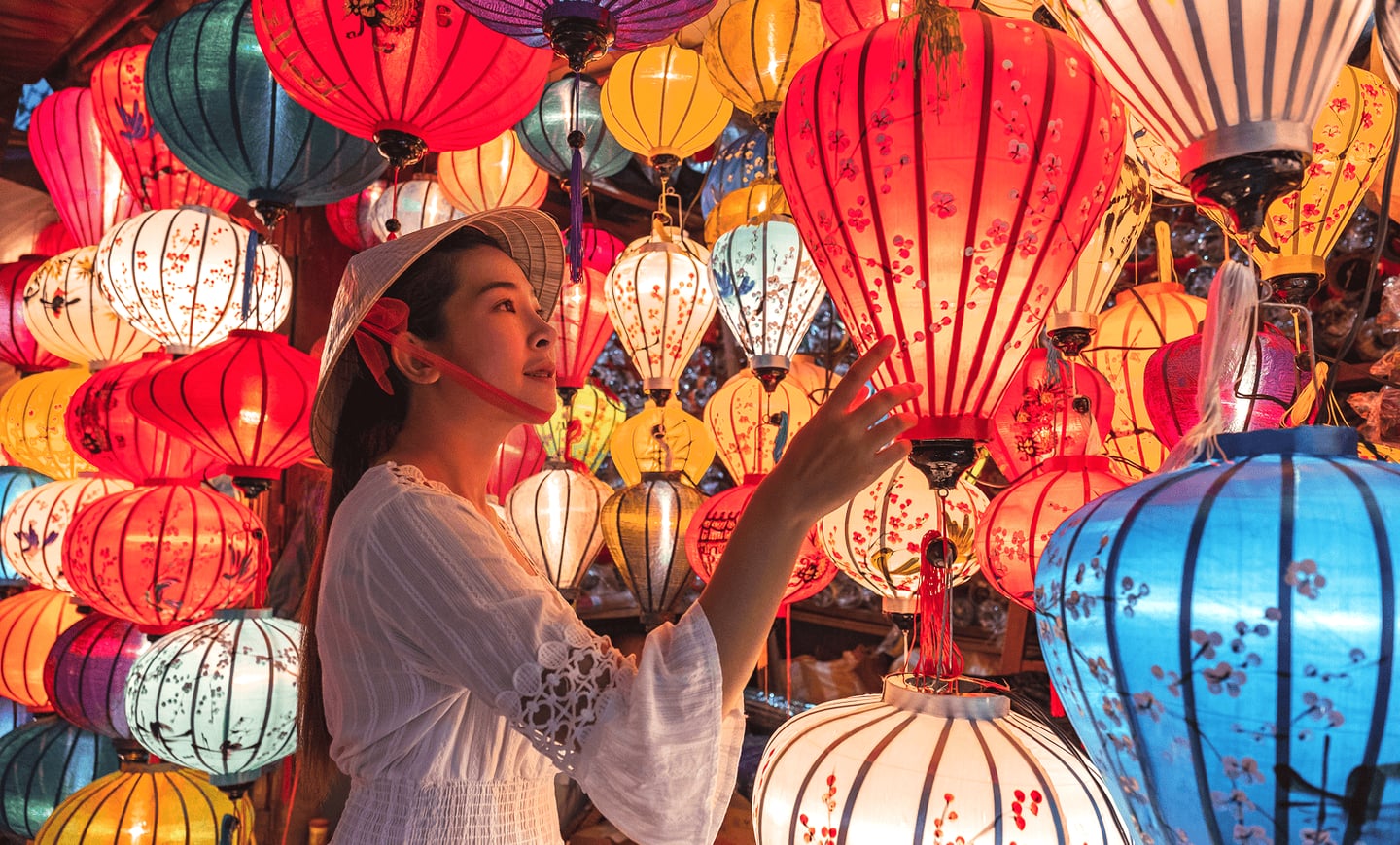 베트남 호이안의 밤거리에서 전통 모자를 쓴 여성이 화려하게 빛나는 형형색색의 수많은 등불들을 배경으로 포즈를 취하고 있는 모습입니다.