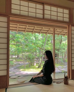 고즈넉한 일본 전통 가옥 다다미에 앉아 창밖으로 보이는 싱그러운 초록빛 정원과 석등을 감상하며 휴식을 취하는 여성의 모습입니다.