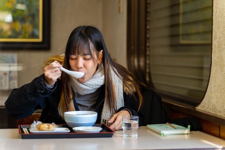 식당 안에서 검은색 코트와 목도리를 두른 여성이 쟁반에 놓인 따뜻한 국물 요리를 숟가락으로 떠먹으며 식사를 즐기고 있습니다.