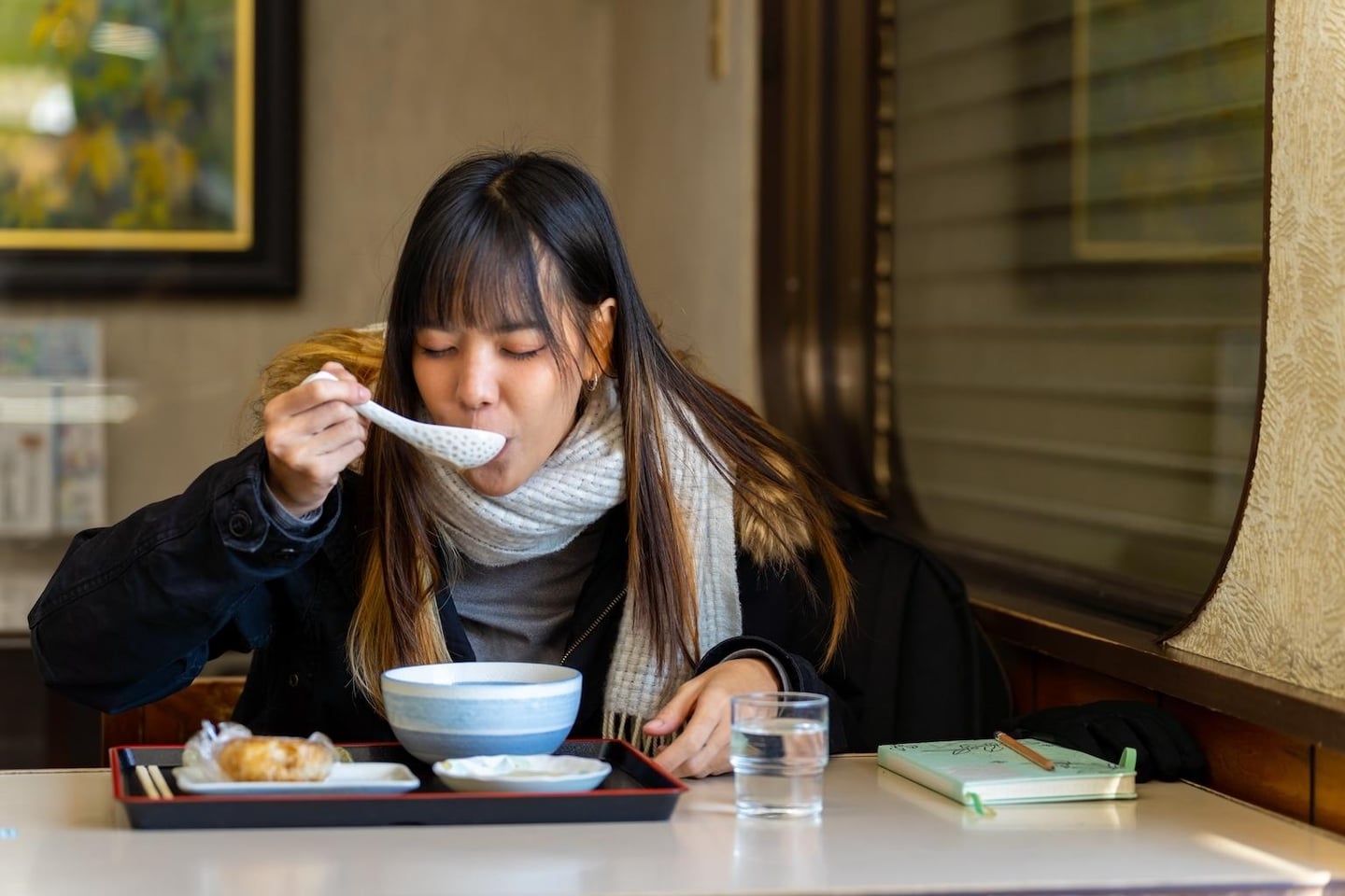 식당 안에서 검은색 코트와 목도리를 두른 여성이 쟁반에 놓인 따뜻한 국물 요리를 숟가락으로 떠먹으며 식사를 즐기고 있습니다.