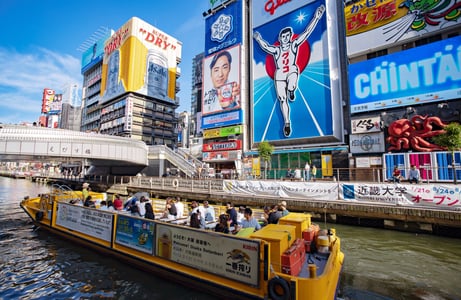 오사카 도톤보리의 상징인 글리코상 전광판과 화려한 빌딩 숲을 배경으로 관광객을 태운 노란색 리버 크루즈가 운하를 따라 유유히 지나가는 풍경입니다.