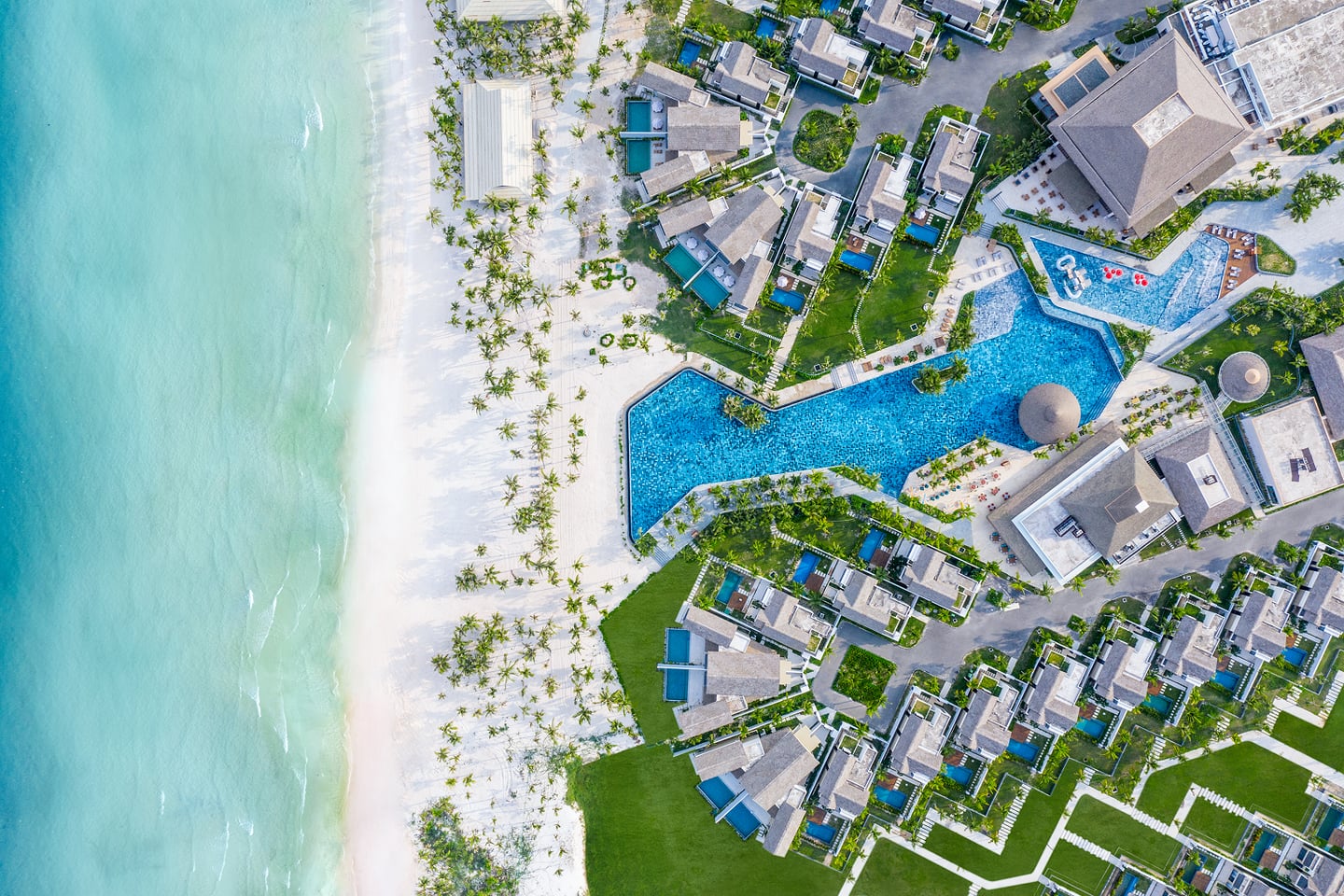 투명한 바다와 백사장이 맞닿은 해안가에 위치한 대규모 럭셔리 리조트의 전경으로, 기하학적인 수영장과 빌라들이 어우러진 이국적인 풍경입니다.