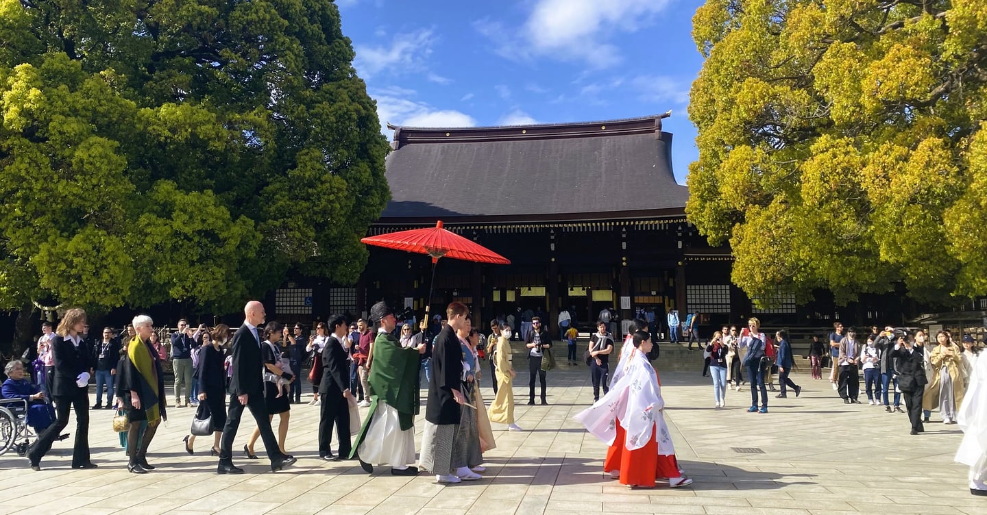 푸른 하늘 아래 일본 전통 사원 광장에서 붉은 양산을 앞세워 전통 의상을 차려입은 사람들이 행렬을 지어 이동하는 모습입니다.