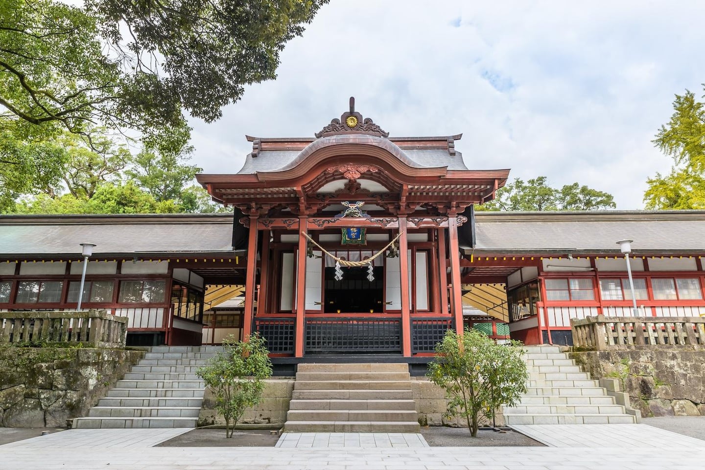 울창한 숲을 배경으로 붉은 목조 구조와 기와지붕이 조화를 이루는 일본 신사의 정면 모습과 석조 계단 풍경입니다.