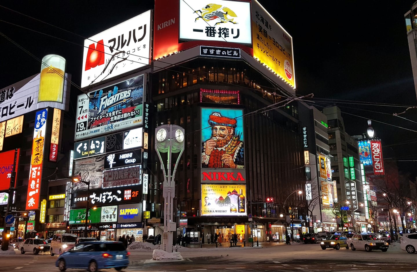 삿포로 스스키노의 상징적인 니카 위스키 광고판과 화려한 네온사인이 빛나는 밤거리에서 자동차들이 눈 쌓인 도로를 주행하는 모습입니다.