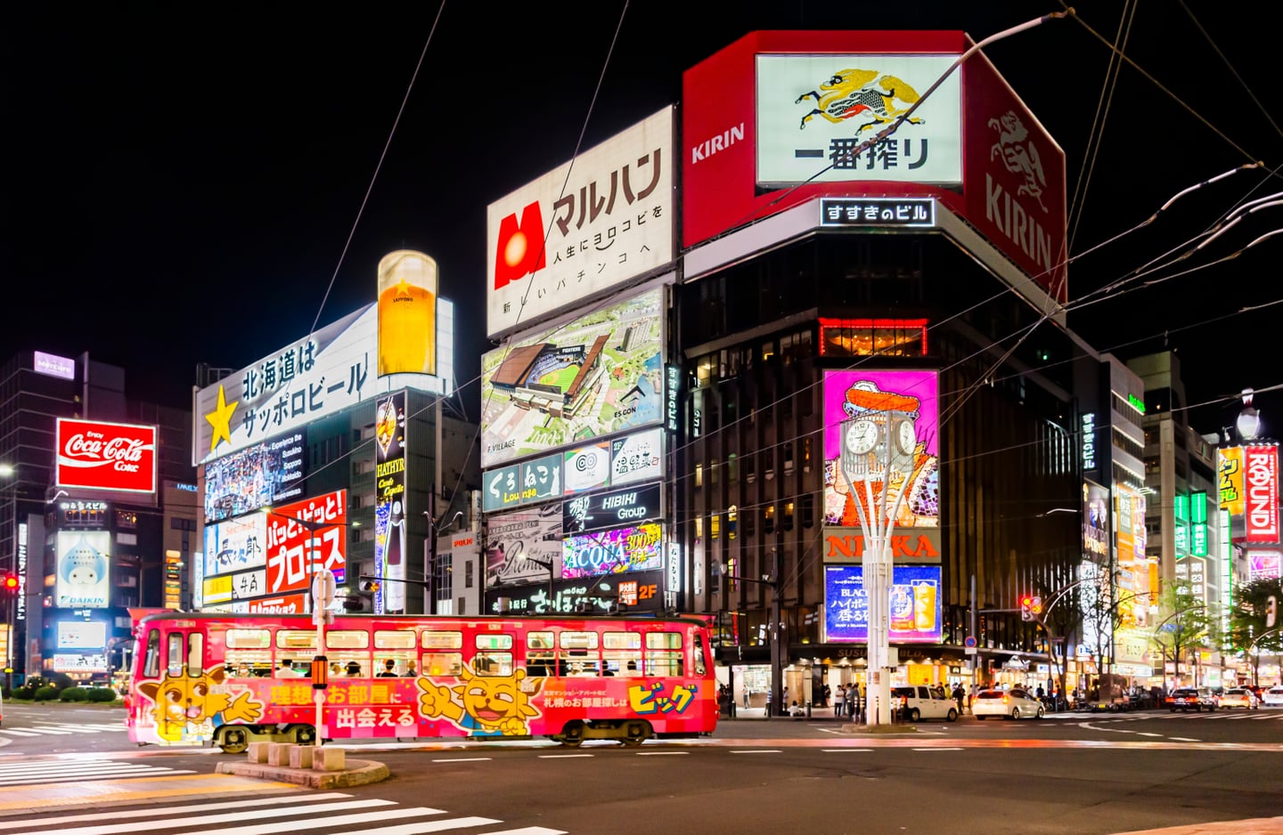 화려한 네온사인 광고판이 가득한 삿포로 스스키노 교차로를 배경으로 분홍색 캐릭터 노면전차가 도로 위를 달리는 역동적인 야경 모습입니다.