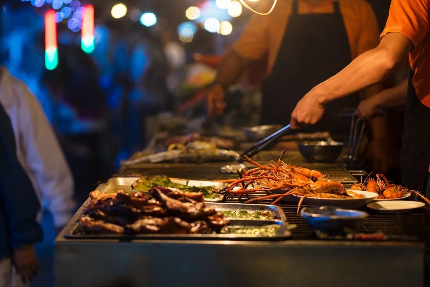 화려한 조명이 빛나는 야시장에서 요리사가 집게를 이용해 그릴 위 신선한 새우와 고기 요리를 정성스럽게 조리하는 장면입니다.