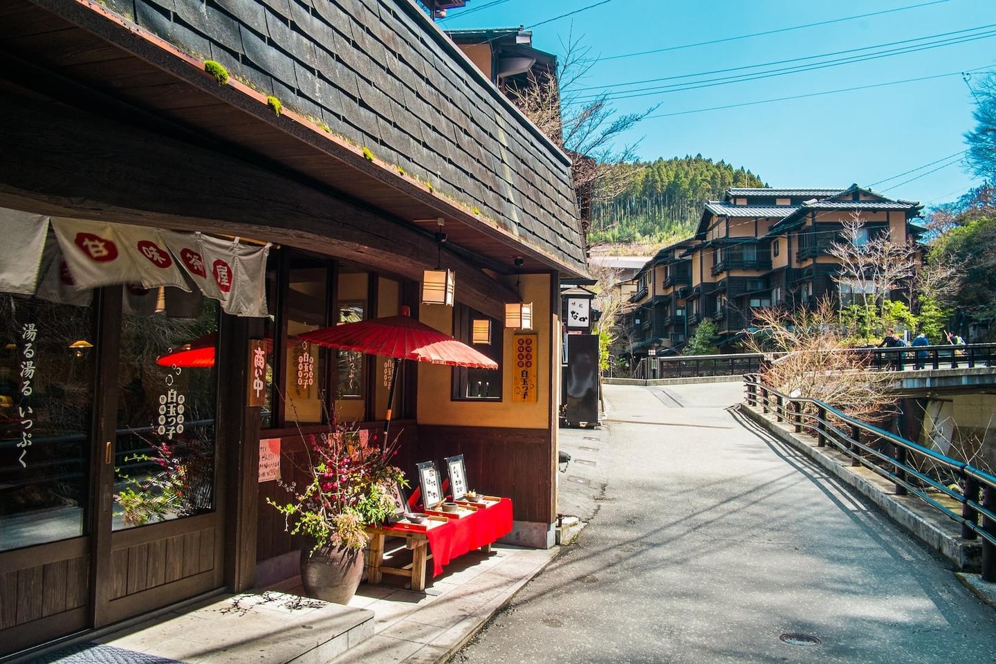 전통적인 목조 가옥과 붉은 파라솔이 돋보이는 일본 온천 마을의 고즈넉한 거리 풍경으로 멀리 산과 다리가 보입니다.