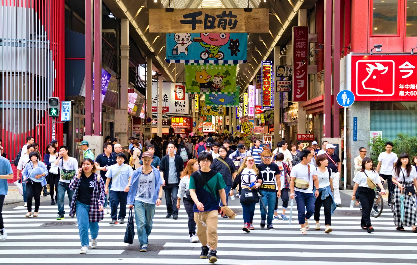 오사카 센니치마에 상점가 입구 횡단보도에서 다양한 차림의 관광객들과 현지인들이 길을 건너며 북적이는 도심의 일상 모습입니다.