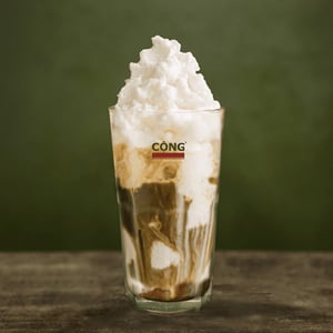 투명한 유리잔에 담긴 진한 갈색 커피와 그 위로 풍성하게 솟아오른 하얀 코코넛 스무디가 대조를 이루는 베트남 스타일의 커피 음료입니다.