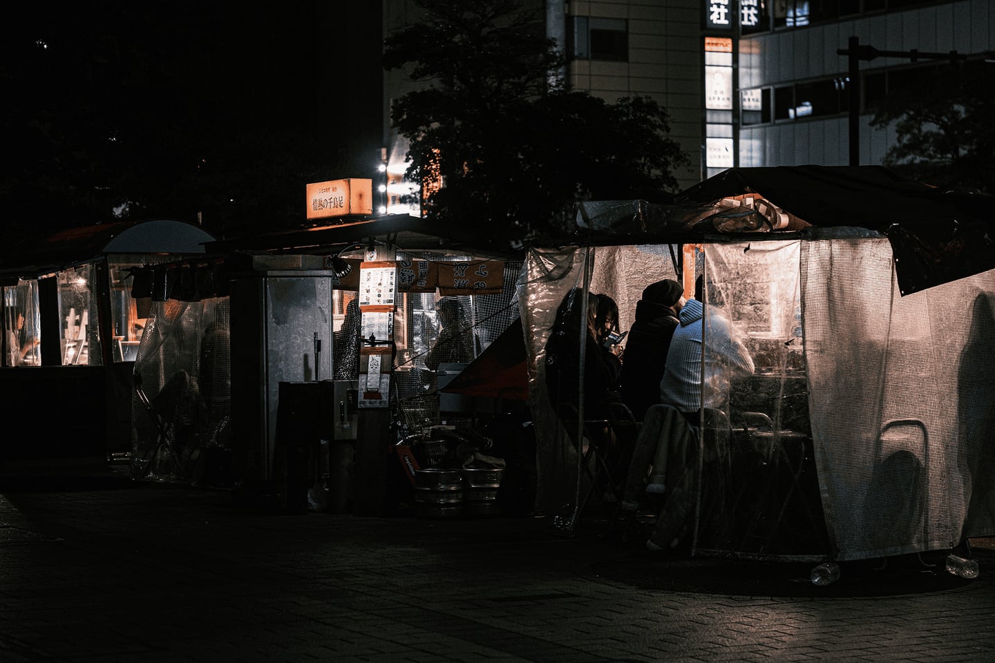 밤의 도심 거리에서 환한 조명이 켜진 비닐 천막 포장마차 안에 사람들이 모여 앉아 야식을 즐기는 아늑하고 서정적인 풍경입니다.