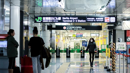 나리타 공항 제2·3터미널역 JR선 입구에서 여행객들이 안내 표지판 아래를 지나가거나 대기하고 있는 실내 전경입니다.