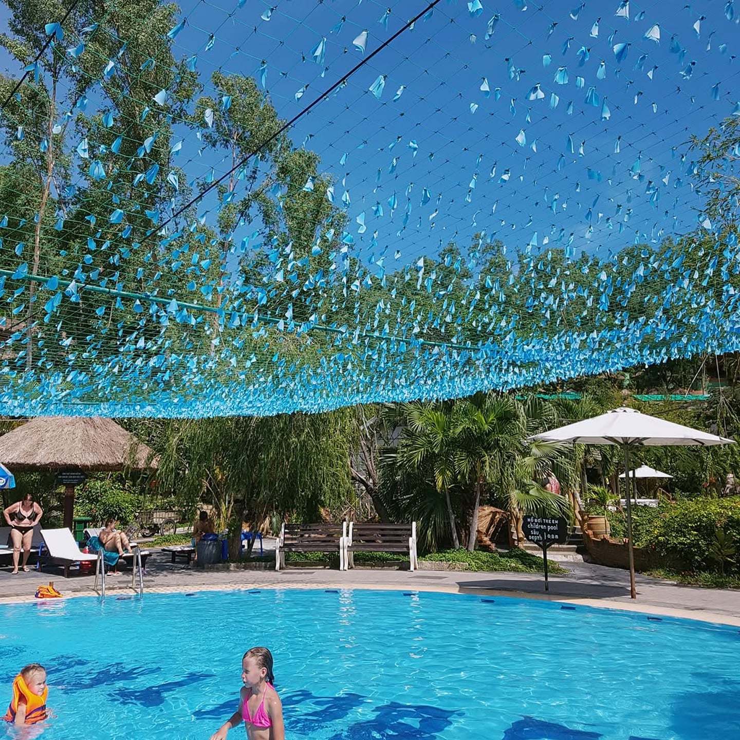 파란 장식물이 가득한 그물 아래 시원한 야외 수영장에서 아이들이 수영을 하고 주변 나무와 파라솔이 어우러진 평화로운 리조트 풍경입니다.