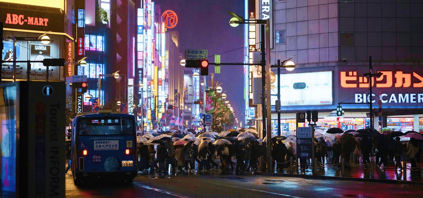 화려한 네온사인이 가득한 도쿄 신주쿠의 밤거리에서 우산을 쓴 인파가 횡단보도를 건너고 있으며 젖은 노면 위로 도시의 불빛이 아름답게 반사되는 풍경입니다.