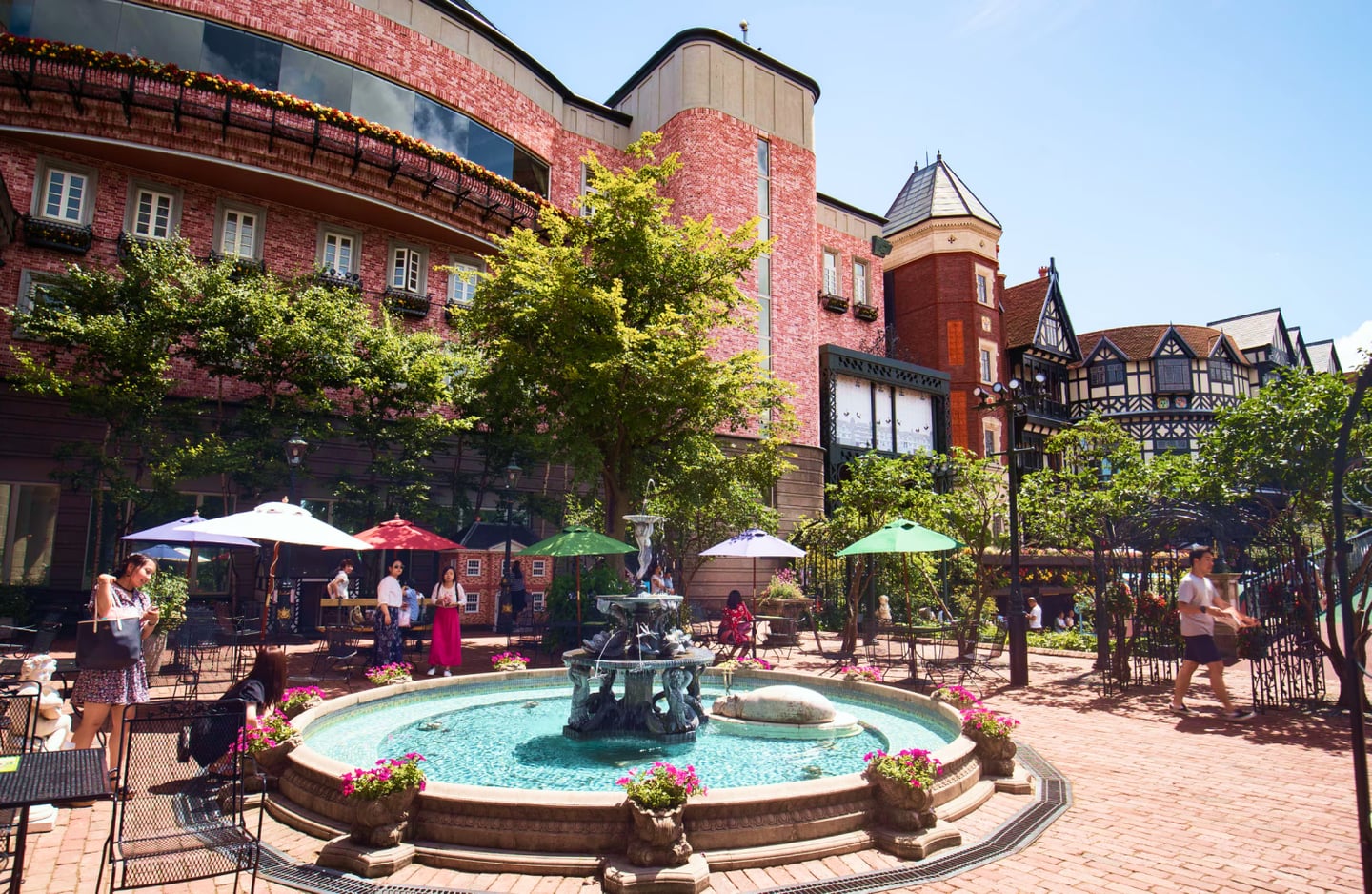 붉은 벽돌 건물과 푸른 나무들이 어우러진 야외 광장에서 시원하게 물을 뿜어내는 원형 분수대와 알록달록한 파라솔 아래 휴식을 취하는 사람들의 모습입니다.