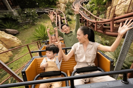 야외 테마파크에서 롤러코스터를 타고 내려오며 두 손을 높이 들고 환호하는 행복한 가족의 역동적인 모습입니다.