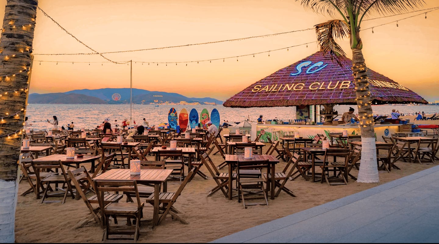 황금빛 노을이 물든 해변가 야외 바에 늘어선 나무 테이블과 의자들 너머로 푸른 바다와 산의 능선이 펼쳐지는 평화로운 저녁 풍경입니다.