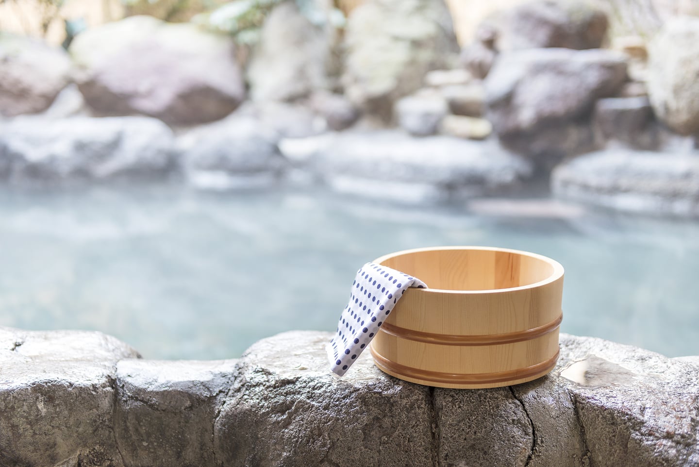 따뜻한 김이 서린 야외 노천탕 바위 위에 놓인 전통적인 나무 대야와 푸른색 점무늬 수건이 평화로운 휴식의 분위기를 전달합니다.