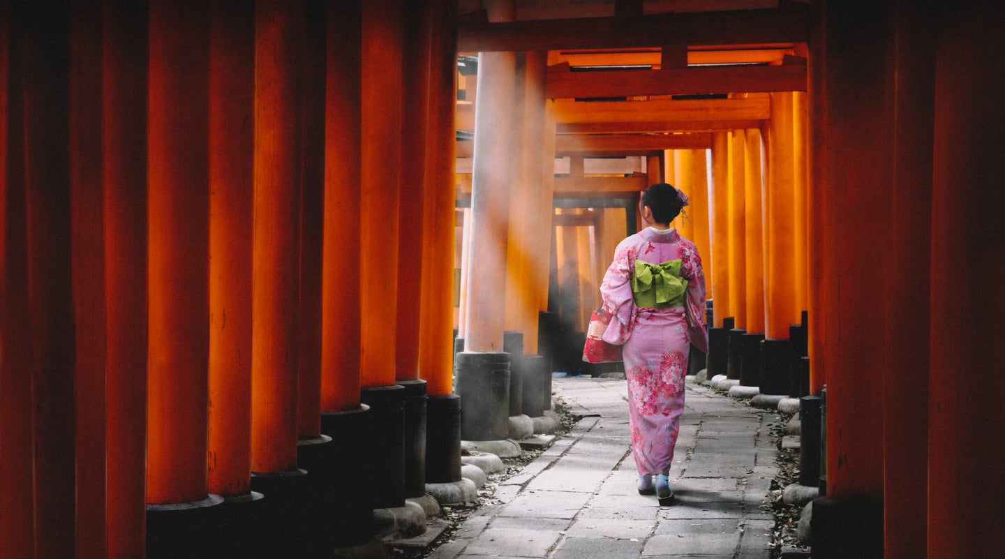일본 교토 후시미 이나리 신사의 끝없이 이어진 붉은 도리이 길을 따라 화사한 분홍색 기모노를 입은 여성이 햇살을 받으며 걸어가는 뒷모습입니다.