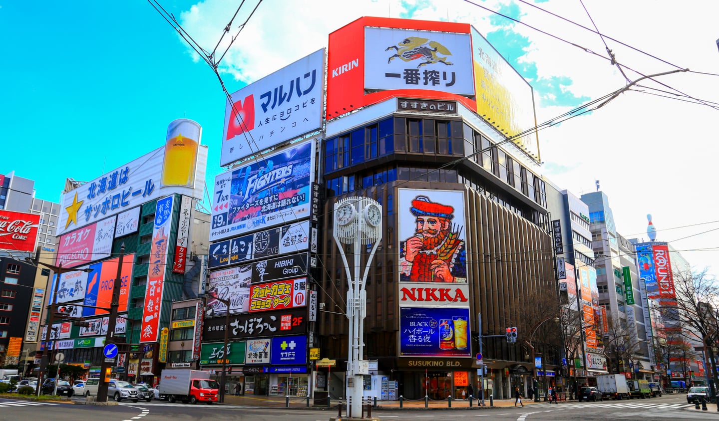 삿포로 스스키노의 랜드마크인 니카 위스키 광고판과 기린 맥주 등 화려한 간판들이 가득한 도심 빌딩 숲의 낮 풍경입니다.