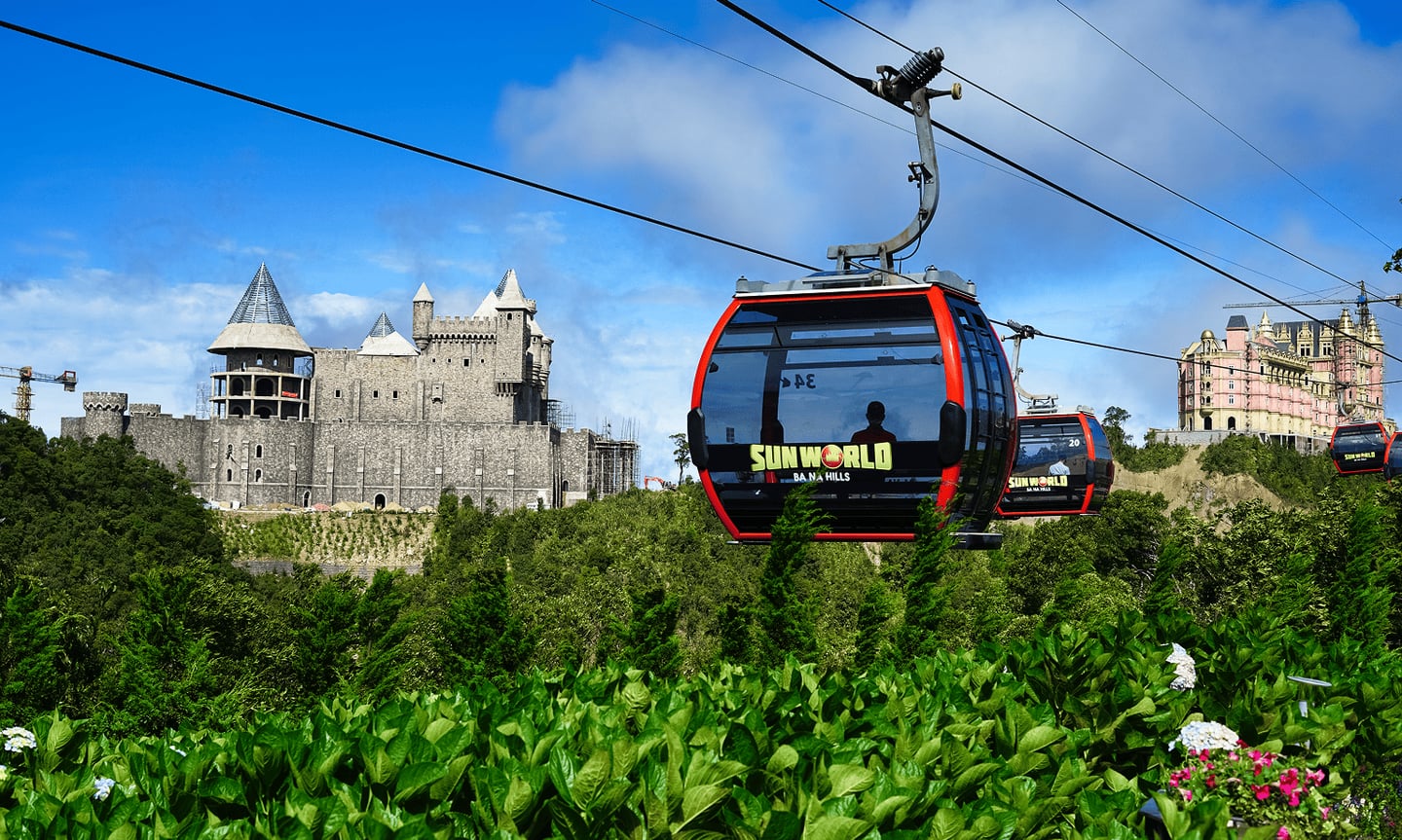다낭 바나힐의 푸른 숲과 고성 스타일 건물을 배경으로 공중에 떠 있는 빨간색 썬월드 케이블카의 평화로운 풍경입니다.