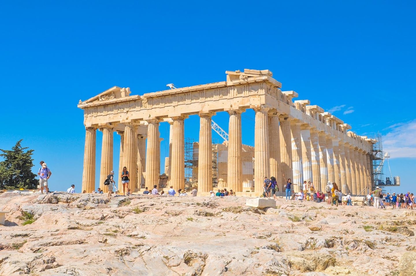 쾌청한 날씨 속에서 고대 그리스의 상징인 파르테논 신전의 거대한 기둥들과 그 주변을 구경하는 수많은 여행객들의 활기찬 모습입니다.