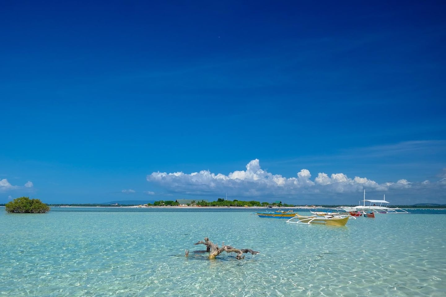투명한 바다 위에 떠 있는 알록달록한 방카 보트들과 수평선 너머로 보이는 푸른 섬, 그리고 맑고 높은 하늘이 어우러진 평화로운 휴양지 풍경입니다.