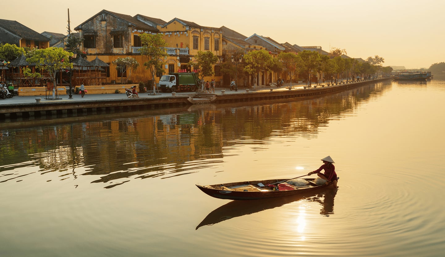 황금빛 노을이 내리는 베트남 호이안 투본강에서 전통 모자 농라를 쓴 어부가 작은 목선을 타고 잔잔한 물결 위를 지나가는 평화로운 풍경입니다.