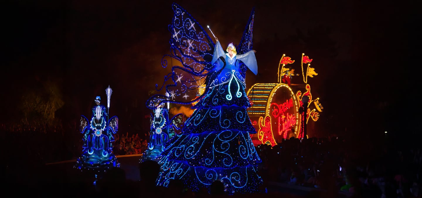 밤하늘을 배경으로 푸른 빛의 화려한 전구 장식이 돋보이는 요정 캐릭터와 퍼레이드 행렬이 테마파크의 야간 축제 분위기를 자아냅니다.
