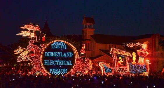 밤하늘 아래 화려한 조명으로 빛나는 도쿄 디즈니랜드 일렉트리컬 퍼레이드 차량과 이를 구경하는 수많은 인파의 모습입니다.
