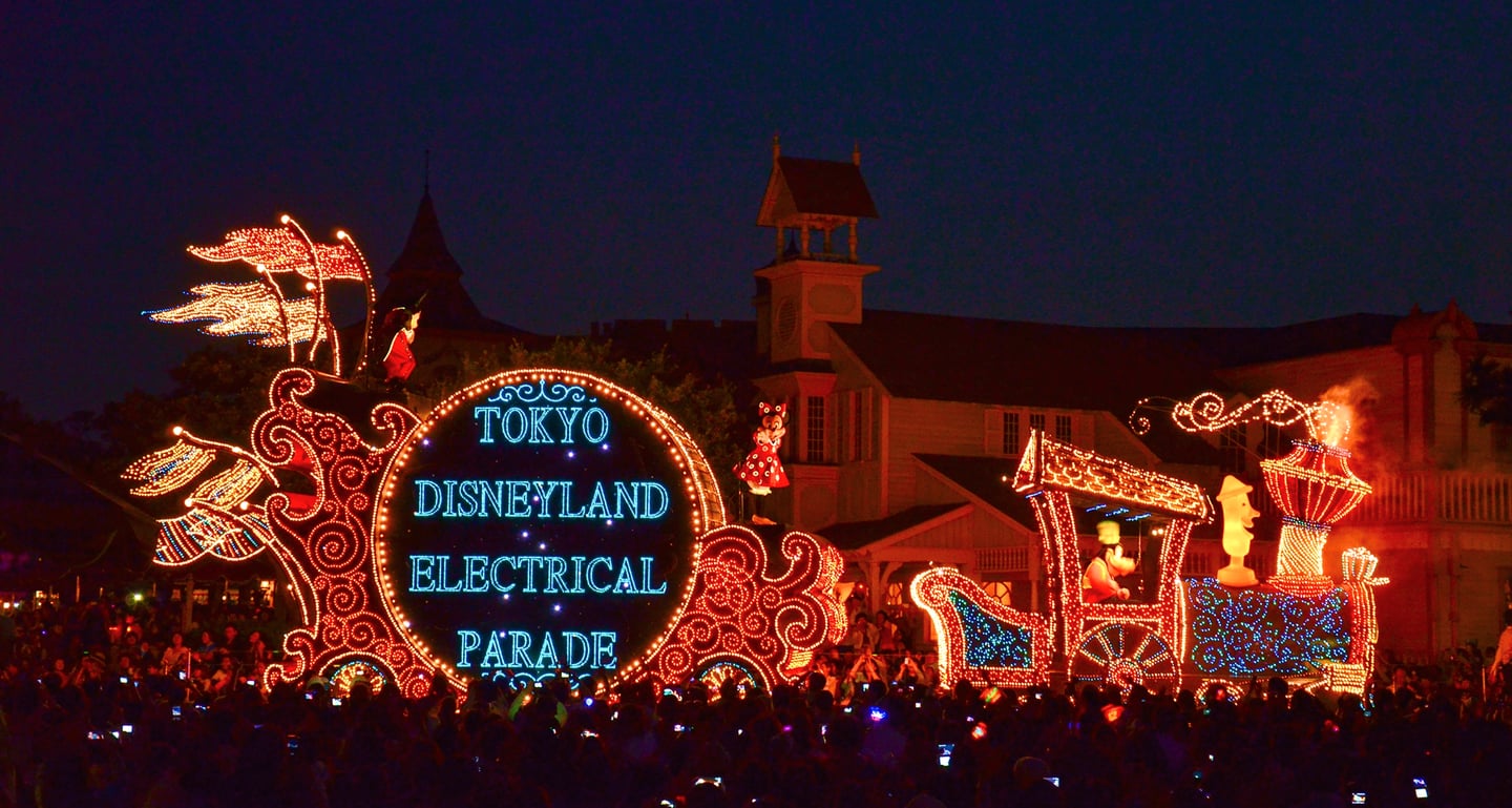 밤하늘 아래 화려한 조명으로 빛나는 도쿄 디즈니랜드 일렉트리컬 퍼레이드 차량과 이를 구경하는 수많은 인파의 모습입니다.