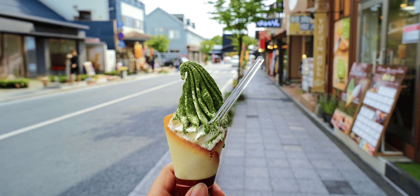 일본 유후인 상점가 거리를 배경으로 랑그드샤 콘에 담긴 먹음직스러운 말차 소프트 아이스크림을 손에 들고 있는 모습입니다.