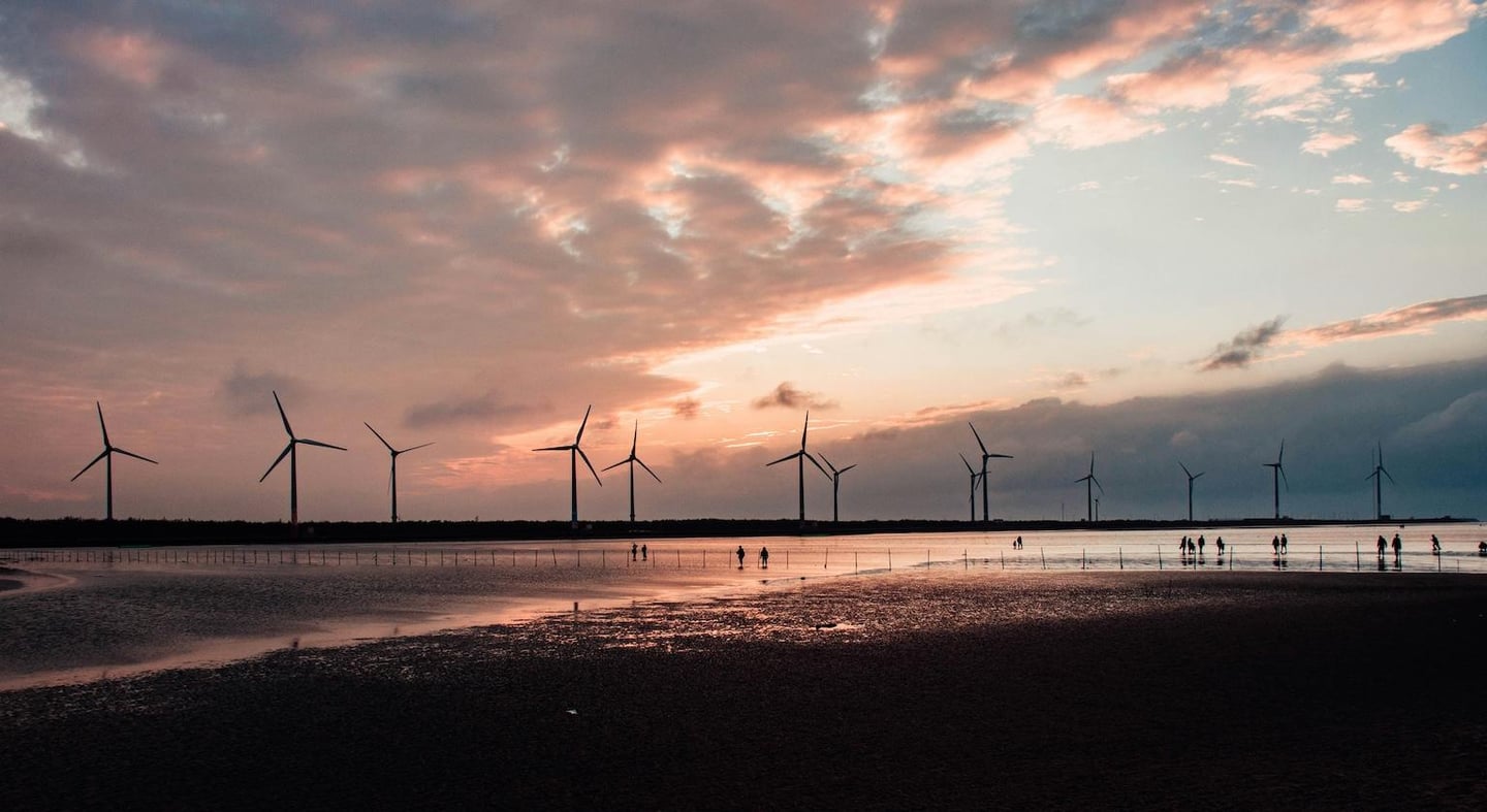 해 질 녘 붉은 노을이 가득한 해안가 갯벌에서 풍력 발전기를 배경으로 여유롭게 산책을 즐기는 사람들의 실루엣이 담긴 풍경입니다.