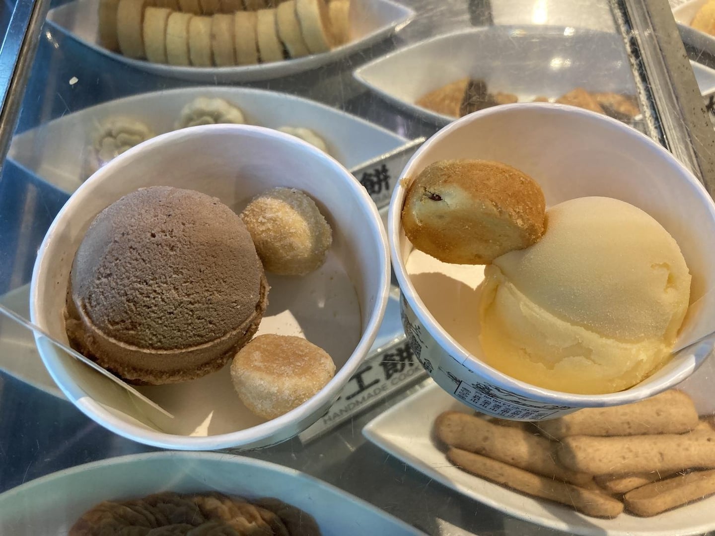 유리 진열장 너머로 보이는 초코와 바닐라 아이스크림 컵 위에 동그란 수제 쿠키가 토핑으로 올라가 있는 달콤한 디저트의 모습입니다.