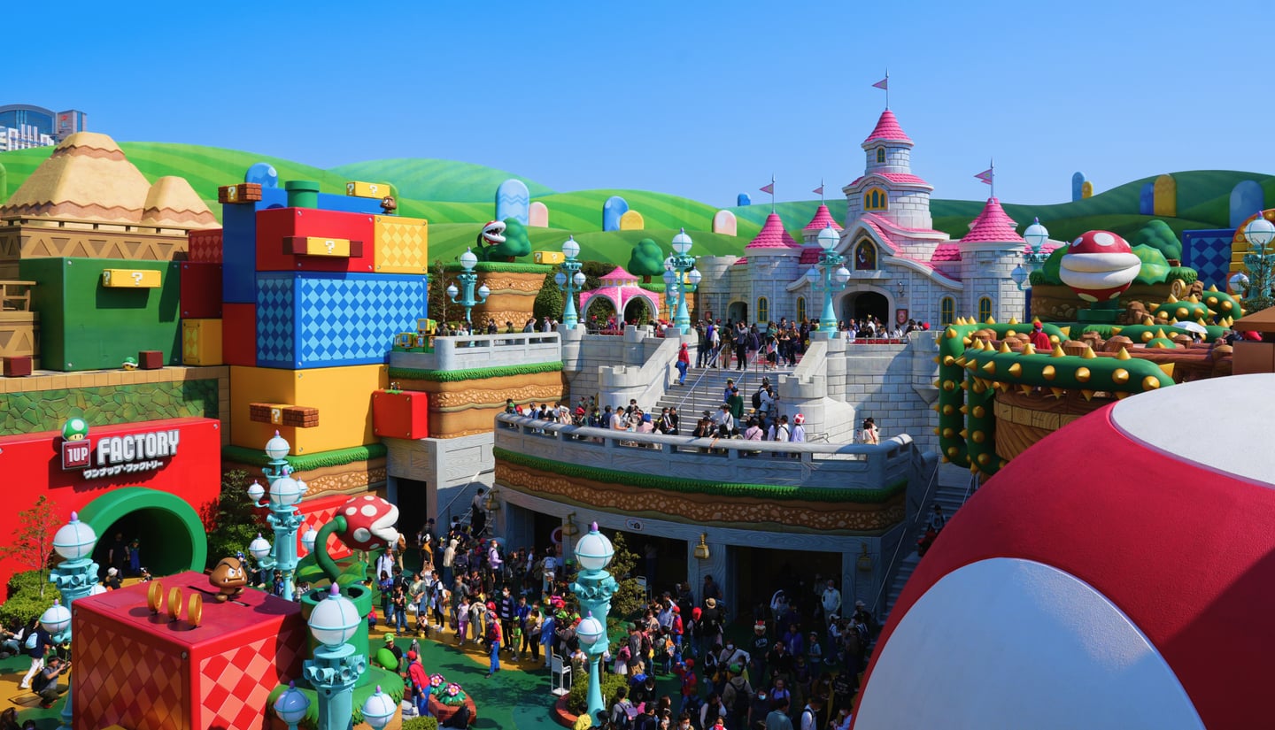 푸른 하늘 아래 펼쳐진 슈퍼 닌텐도 월드의 화려한 성과 블록 건물들 사이로 많은 사람들이 즐겁게 이동하는 테마파크 풍경입니다.