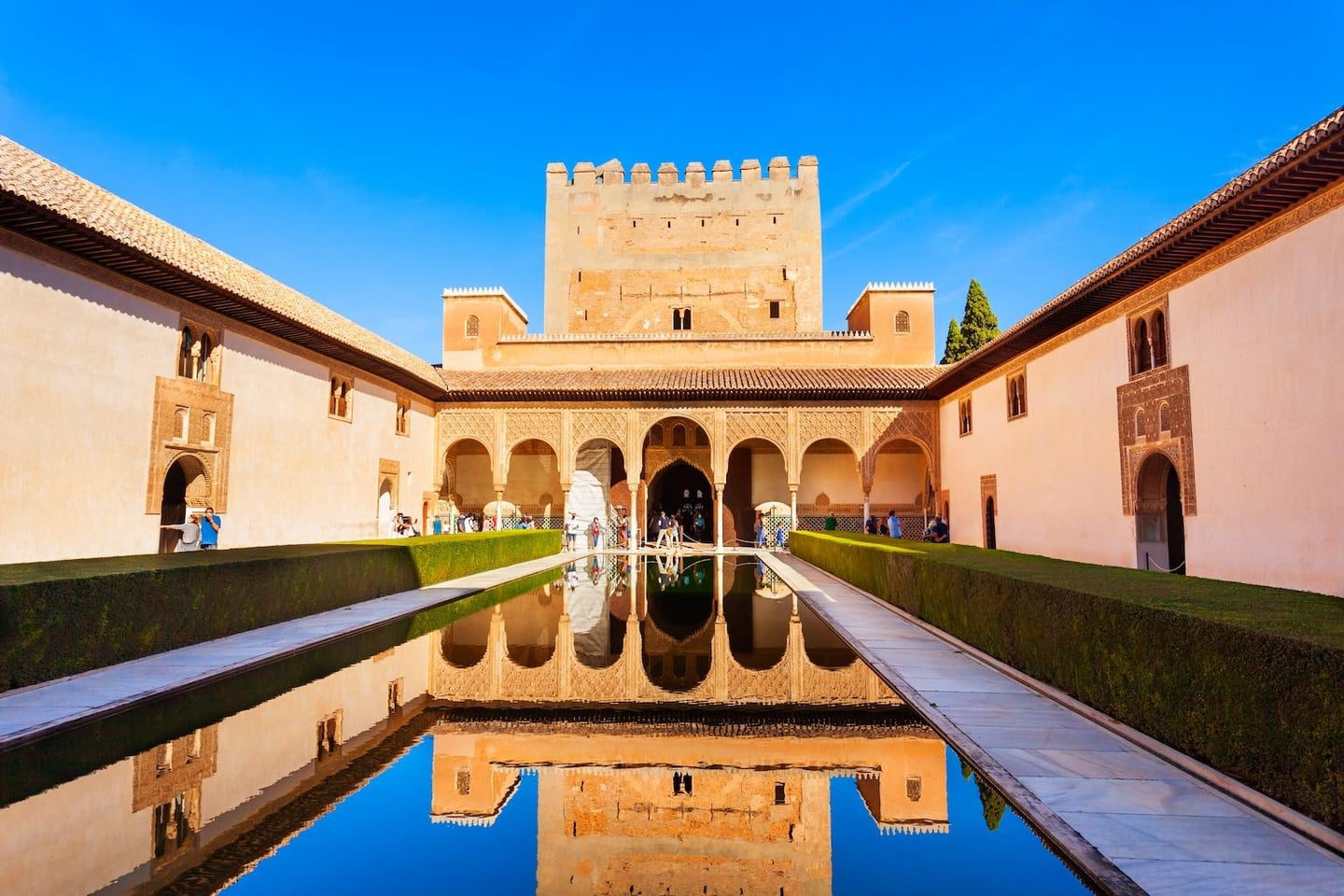 푸른 하늘 아래 알람브라 궁전의 정교한 아치형 건물과 성벽이 중앙 수로의 맑은 물 위로 대칭을 이루며 아름답게 투영된 풍경입니다.