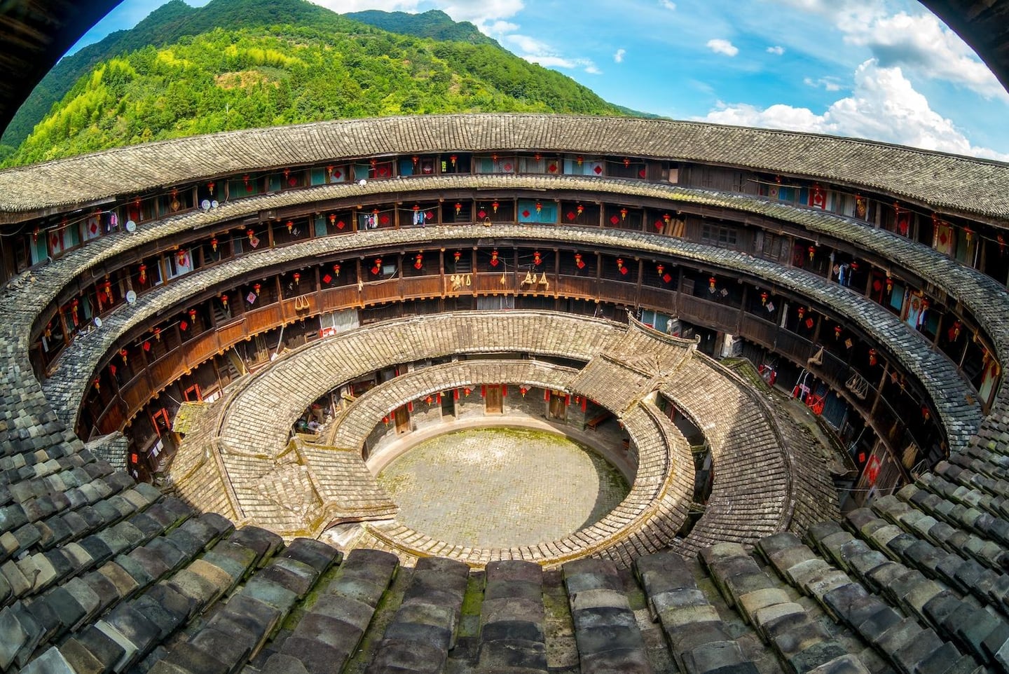 푸른 산을 배경으로 웅장하게 자리 잡은 중국 전통 원형 가옥 토루의 다층 구조와 기와지붕이 내려다보이는 풍경입니다.