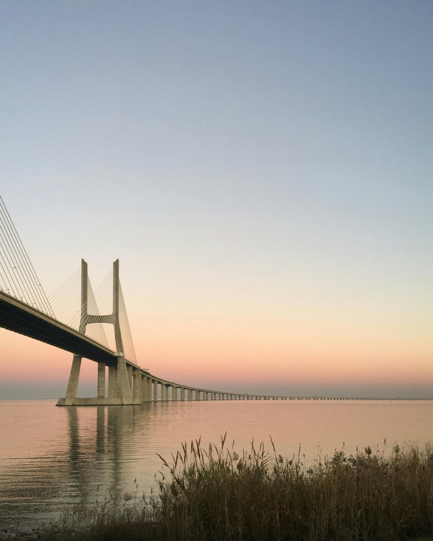 리스본 바스쿠 다 가마 다리, 분홍빛 노을이 물든 하늘 아래 타구스 강 위로 길게 뻗은 현대적인 사장교의 웅장한 실루엣.