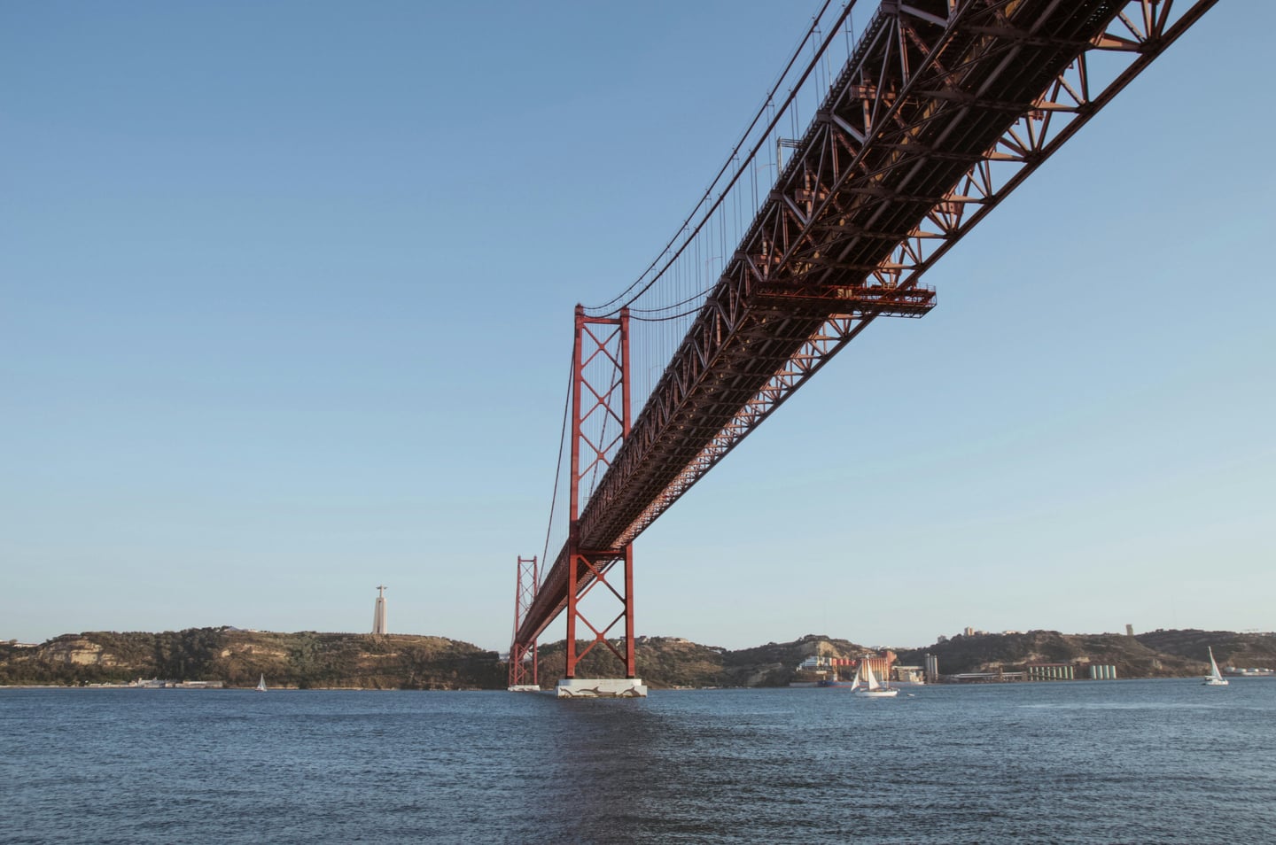 리스본 4월 25일 다리, 푸른 타구스 강 위로 웅장하게 뻗은 붉은색 현수교와 멀리 보이는 그리스도 상의 전경입니다.