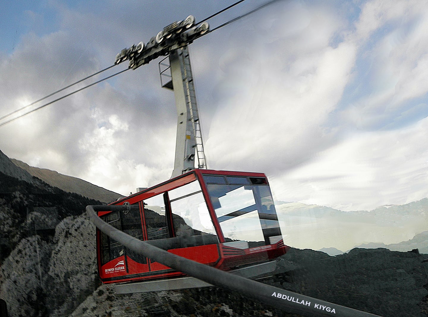안탈리아 올림포스산 케이블카, 웅장한 타흐탈리 산맥의 암벽 위를 가로지르며 구름 사이로 높이 솟아오르는 빨간색 캐빈의 역동적인 풍경입니다.