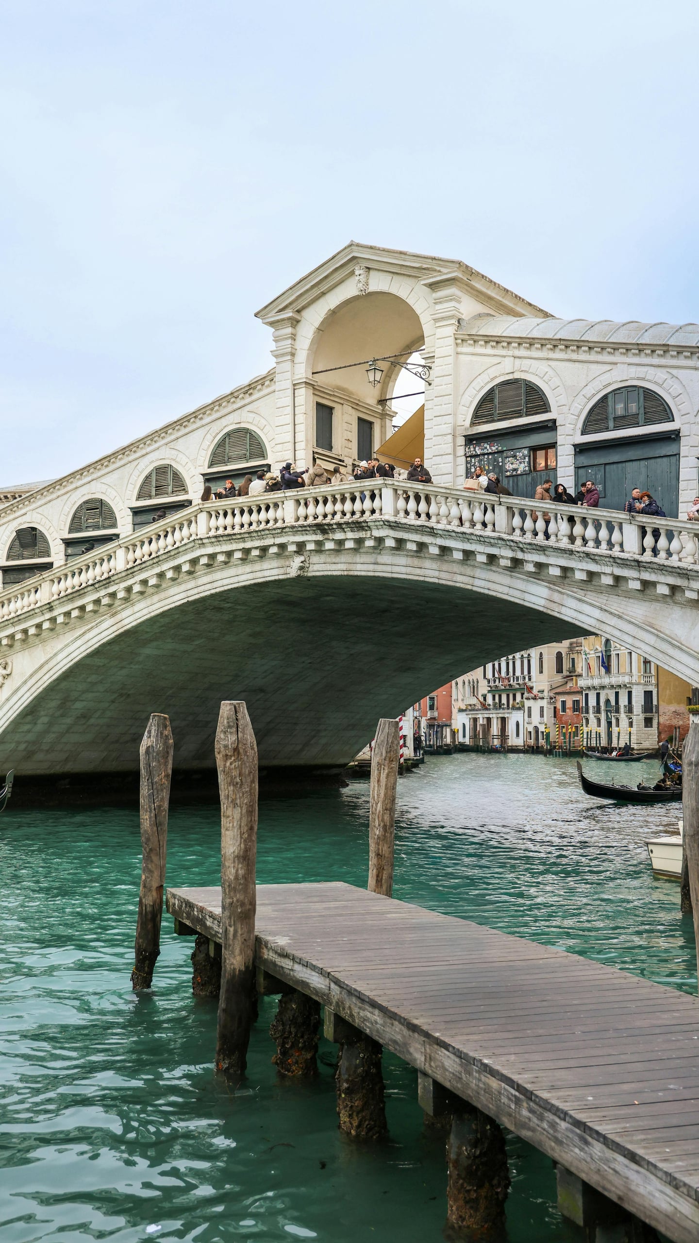 베니스 리알토 다리, 에메랄드빛 운하 위로 웅장하게 솟아오른 흰색 대리석 아치와 그 위를 가득 메운 관광객들의 활기찬 풍경입니다.