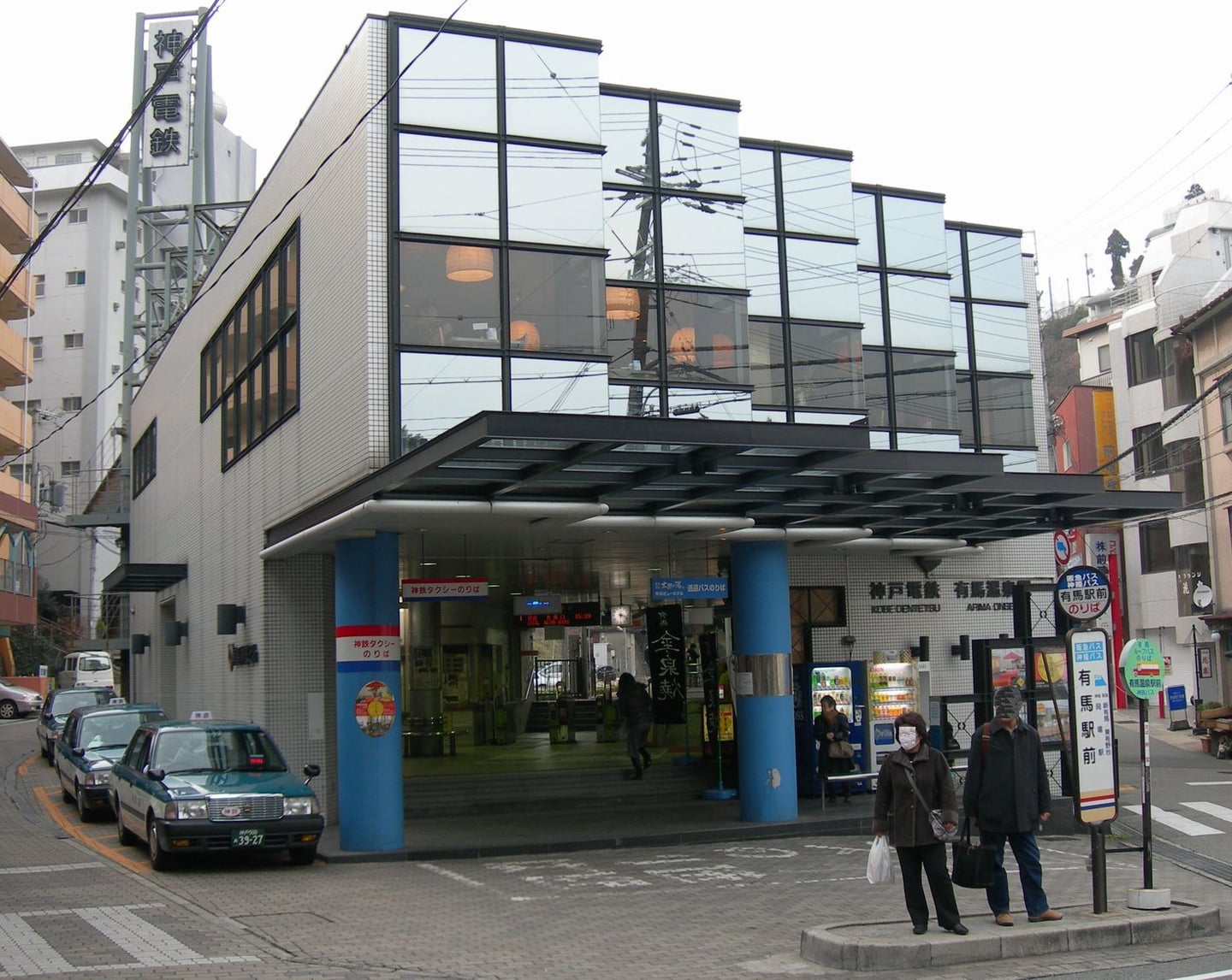 고베 아리마 온천, 현대적인 유리 외벽이 돋보이는 아리마 온천역 건물과 정류장에서 버스를 기다리는 사람들의 일상적인 풍경입니다.