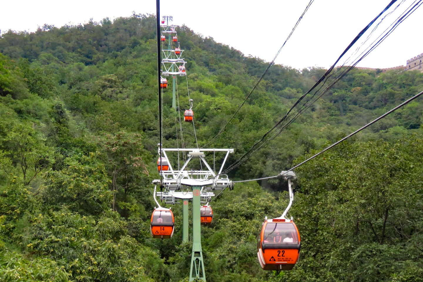 베이징 만리장성 케이블카, 푸른 숲이 우거진 산등성이를 따라 줄지어 이동하는 주황색 곤돌라와 멀리 보이는 성벽의 전경입니다.