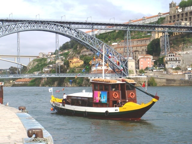 포르투 도루강 크루즈, 웅장한 루이스 1세 다리 아래로 오색 깃발을 단 전통 라벨로 보트가 물결을 가르며 지나가는 평화로운 풍경입니다.