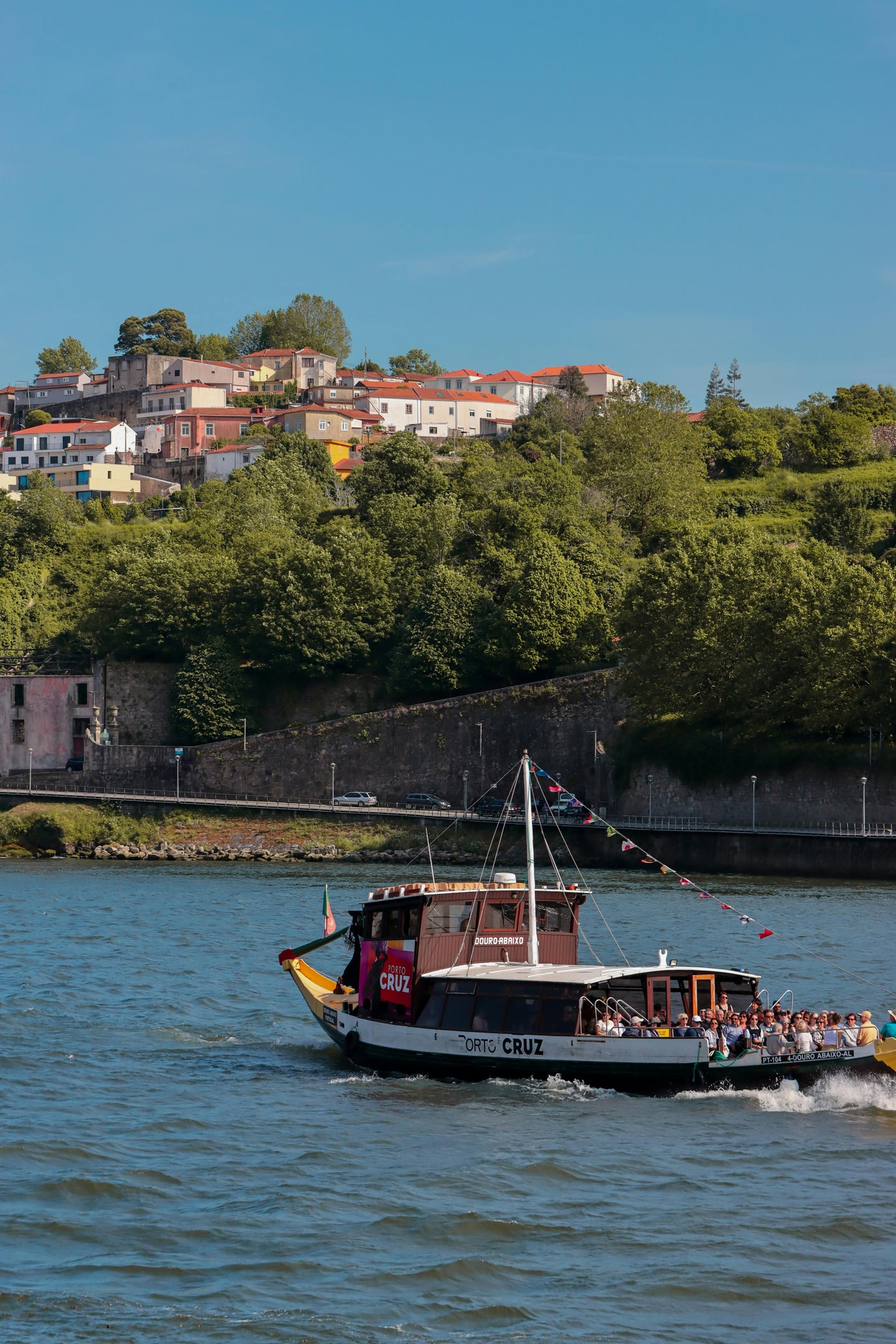 포르투 도루강 크루즈, 푸른 강물 위를 가르며 나아가는 전통 목조 유람선과 언덕 위 붉은 지붕의 마을 풍경이 조화로운 모습입니다.