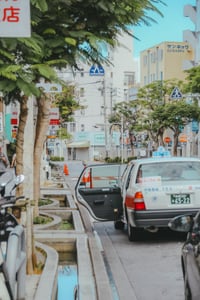 오키나와 국제거리, 푸른 가로수 사이로 문이 열린 택시와 일본어 간판이 늘어선 활기찬 나하 시내의 일상적인 풍경입니다.